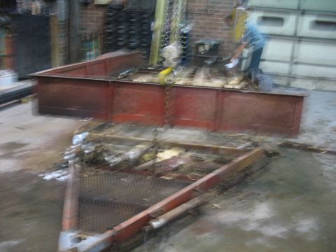 1900 Ford C-350 (Centurion Conversions) - A person works on a trailer frame and bed in a cluttered workshop environment.