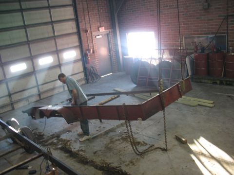 1900 Ford C-350 (Centurion Conversions) - A man is working on a red trailer frame that is suspended by chains inside a garage with large bay doors.
