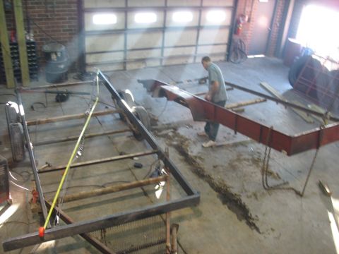 1900 Ford C-350 (Centurion Conversions) - A man works on a metal trailer frame in a garage with large overhead doors.