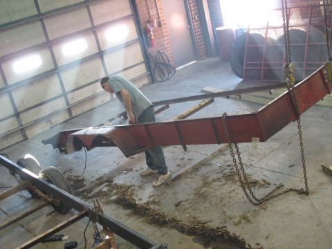 1900 Ford C-350 (Centurion Conversions) - A person examines a red trailer frame suspended by chains inside a workshop.