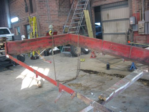 1900 Ford C-350 (Centurion Conversions) - A red metal trailer frame is suspended by chains inside a workshop.