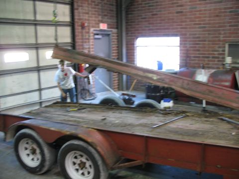 1900 Ford C-350 (Centurion Conversions) - A person uses a crane to load a metal beam onto a trailer inside a garage.