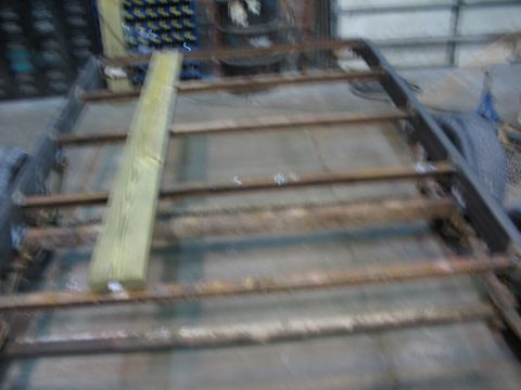 1900 Ford C-350 (Centurion Conversions) - Close-up of a rusty metal frame structure with a wooden plank on top, in a workshop setting.