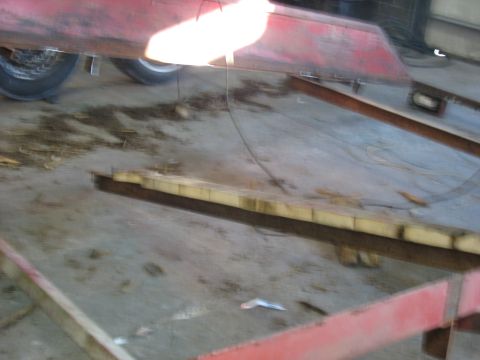 1900 Ford C-350 (Centurion Conversions) - A red metal trailer frame with yellow padding and a tire is shown in a workshop setting.