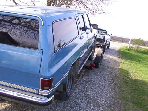 1900 Ford C-350 (Centurion Conversions) - A blue SUV is loaded onto a trailer, with a white pickup truck visible behind it on a gravel road.