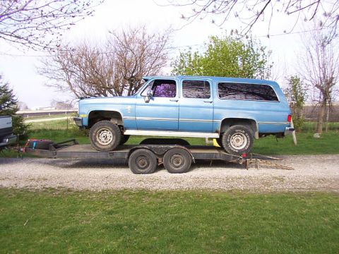 1900 Ford C-350 (Centurion Conversions) - A blue extended cab pickup truck is transported on a dual-axle trailer, with trees and grass in the background.