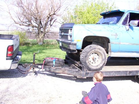 1900 Ford C-350 (Centurion Conversions) - A blue pickup truck is loaded onto a trailer, with a white truck visible in the background.