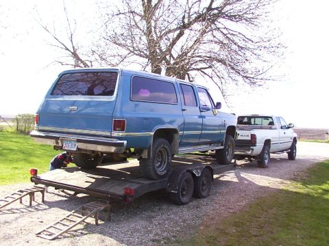 1900 Ford C-350 (Centurion Conversions) - A blue SUV is loaded onto a trailer, with a white pickup truck parked behind it on a gravel surface.