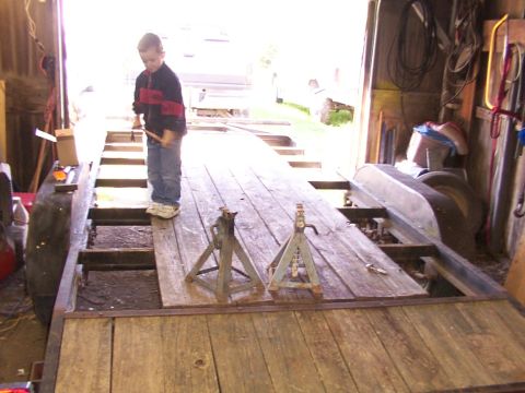 1900 Ford C-350 (Centurion Conversions) - A boy stands on the wooden deck of a trailer frame supported by jack stands in a garage.