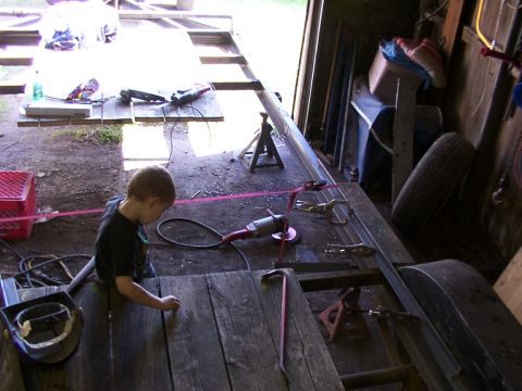 1900 Ford C-350 (Centurion Conversions) - Child works on trailer frame construction in a workshop setting with tools and equipment.