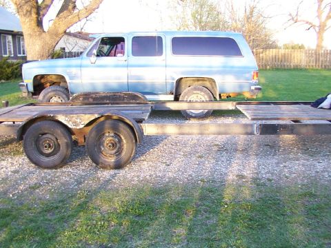 1900 Ford C-350 (Centurion Conversions) - A light blue SUV is loaded onto a trailer, parked on a gravel surface with grass and a fence in the background.