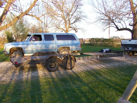 1900 Ford C-350 (Centurion Conversions) - A blue Chevrolet Suburban is parked next to a trailer, with a Dodge pickup truck visible in the background.