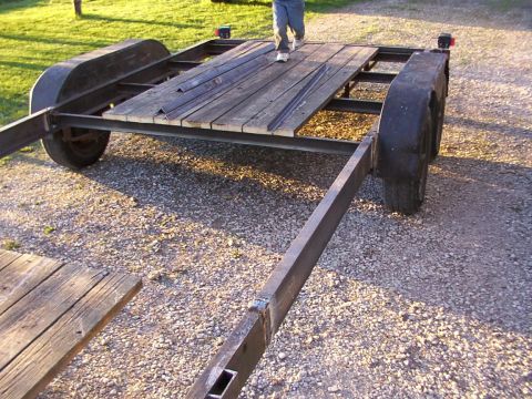 1900 Ford C-350 (Centurion Conversions) - A dual axle trailer with a wooden deck is shown from a low angle, parked on a gravel surface.