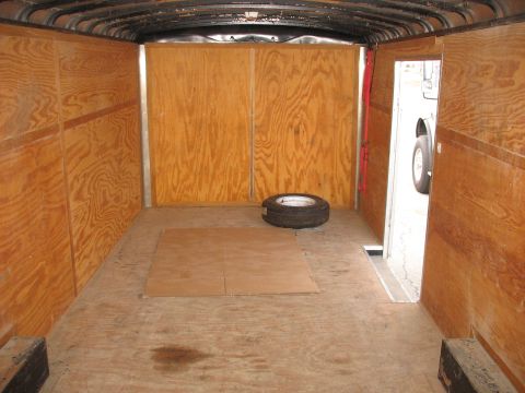 1900 Ford C-350 (Centurion Conversions) - The interior of an empty cargo trailer shows wooden paneling and a spare tire, with a glimpse of a vehicle outside.