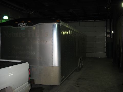 1900 Ford C-350 (Centurion Conversions) - An enclosed trailer with 'Nature Hills Nursery' branding is parked next to a pickup truck inside a garage.