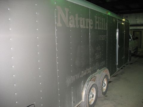 1900 Ford C-350 (Centurion Conversions) - A silver enclosed trailer with "Nature Hills Nursery" and contact information is parked indoors next to a truck.