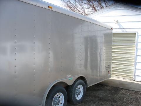 1900 Ford C-350 (Centurion Conversions) - A silver enclosed cargo trailer is parked next to a white garage with a roll-up door. Some text is visible on the trailer's side.