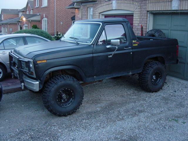 Lifted flat black Ford Bronco with rear top removed, big mud tires and brush guard, parked on gravel driveway