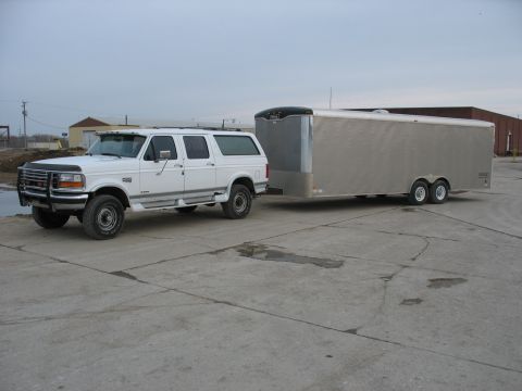 1900 Ford C-350 (Centurion Conversions) - A white Ford SUV from the 1990s tows a large silver cargo trailer on a paved lot under an overcast sky.