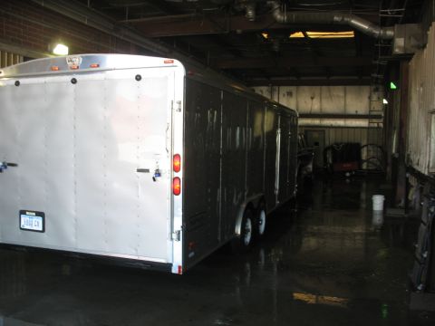 1900 Ford C-350 (Centurion Conversions) - Rear view of a silver enclosed trailer with dual axles, parked on a wet floor inside a building.