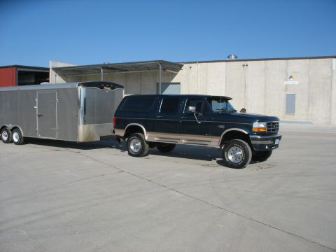 1900 Ford C-350 (Centurion Conversions) - A dark green SUV tows a silver trailer in a paved lot with industrial buildings in the background.