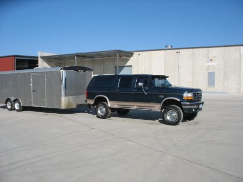 1900 Ford C-350 (Centurion Conversions) - A dark green SUV with tan trim tows a silver trailer in an industrial area under a clear blue sky.