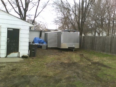 1900 Ford C-350 (Centurion Conversions) - An enclosed cargo trailer sits in a grassy yard next to a white building with a black door.