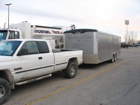 1900 Ford C-350 (Centurion Conversions) - A white pickup truck is parked next to a large silver trailer, with other vehicles and equipment in the background.