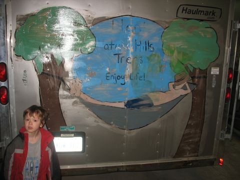 1900 Ford C-350 (Centurion Conversions) - A child stands next to a trailer decorated with a mural depicting a hammock scene and the words 'Hop at Hills Trees Enjoy Life!'.