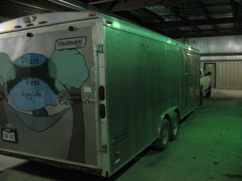 1900 Ford C-350 (Centurion Conversions) - A silver Haulmark trailer with a mural is parked next to a white pickup truck in a dimly lit garage.