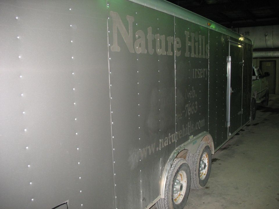 Silver enclosed trailer with 'Nature Hills Nursery' text; a light-colored vehicle is visible in the background
