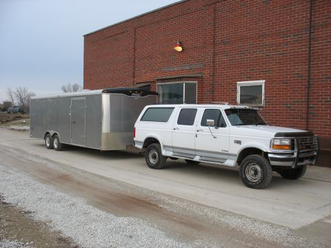1900 Ford C-350 (Centurion Conversions) - White Ford SUV towing a silver trailer parked next to a brick building.