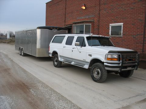 1900 Ford C-350 (Centurion Conversions) - A white Ford Bronco SUV is parked next to a brick building, towing a large silver cargo trailer.