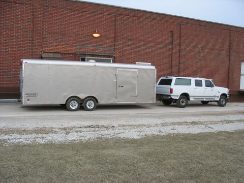 1900 Ford C-350 (Centurion Conversions) - White SUV towing a silver enclosed trailer is parked outside a brick building.
