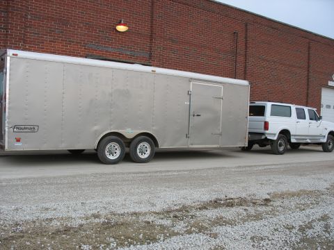 1900 Ford C-350 (Centurion Conversions) - A white SUV is hitched to a large silver enclosed trailer, parked on a gravel surface in front of a brick building.