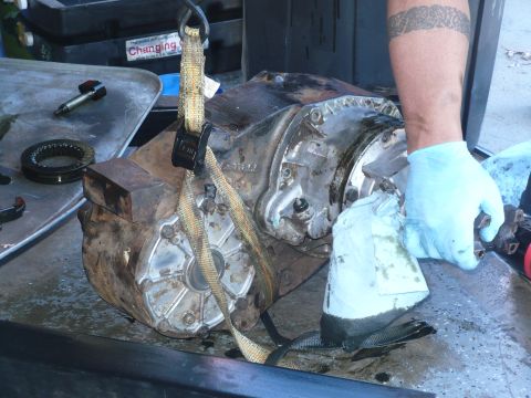 1979 Ford Bronco - A greasy cast-aluminum and cast-iron transfer case sitting on a steel workbench, held by a yellow ratchet strap, with a tattooed forearm in…