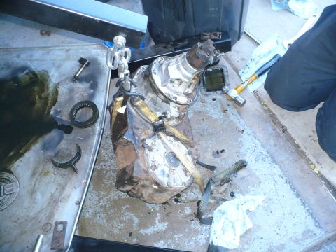 1979 Ford Bronco - A rusty and oil-stained transfer case slung from a hoist chain with a yellow ratchet strap, standing on a shop floor beside a drain pan, sp…