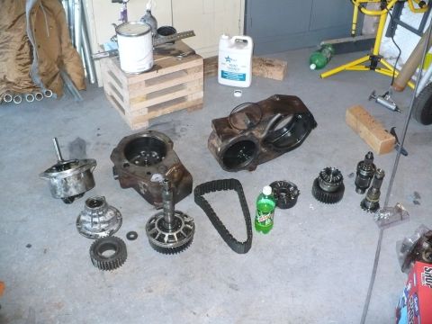 1979 Ford Bronco - A chain-drive transfer case fully disassembled and laid out on a garage floor, showing the split aluminum case halves, oil-stained cast hou…