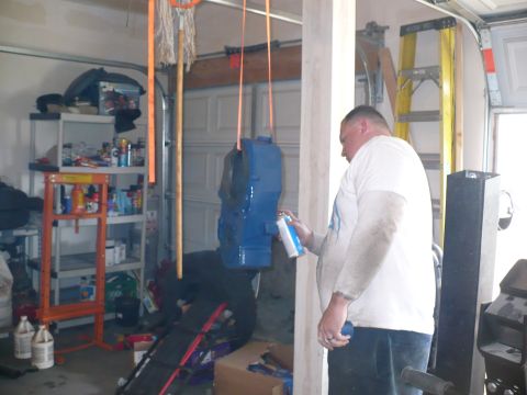 1979 Ford Bronco - Man spray painting a blue part in a garage setting with tools and equipment around.