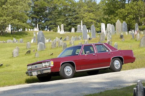 2001 Ford Crown Victoria - A red 2001 Ford Crown Victoria sedan is seen from the side, parked on a road within a cemetery.