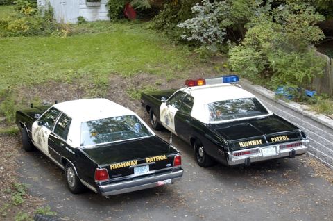 2001 Ford Crown Victoria - Two highway patrol cars are parked side-by-side on a gravel driveway, with trees and a building in the background.