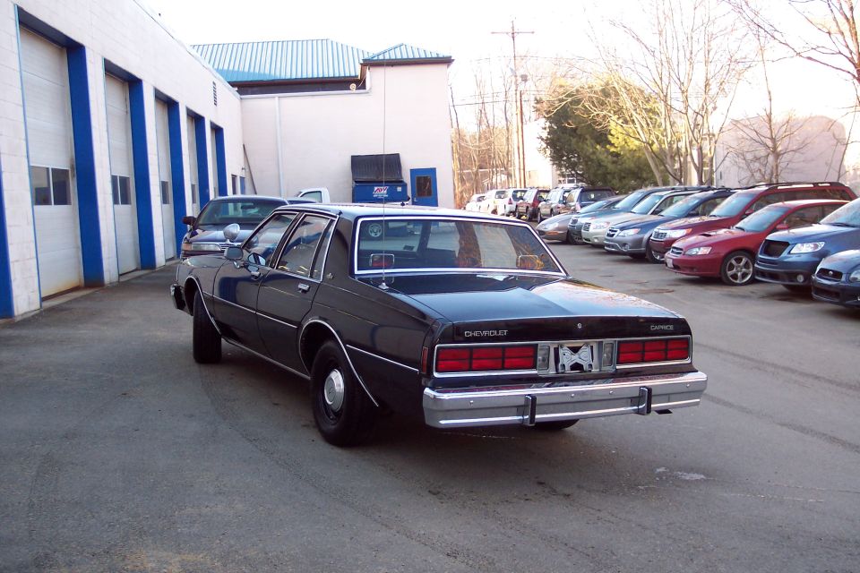 Black Chevrolet Caprice sedan, rear 3/4 view, parked in lot with other cars. Chrome bumper, red taillights