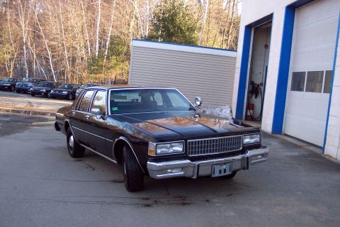 2001 Ford Crown Victoria - A dark-colored sedan, likely a Chevrolet Caprice from the 1980s, is parked in front of a commercial building.
