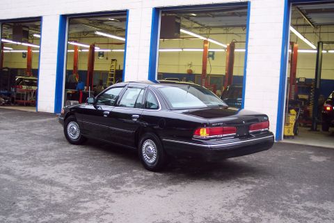 2001 Ford Crown Victoria - This black 2001 Ford Crown Victoria is shown from the rear three-quarter angle, parked in front of garage service bays.