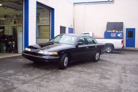 2001 Ford Crown Victoria - A black sedan is parked in front of an open garage bay, with a pickup truck and dumpster nearby.