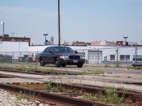 2001 Ford Crown Victoria - A dark sedan, identified as a 2001 Ford Crown Victoria, is seen from a 3/4 front angle in an industrial area with train tracks in the foreg…