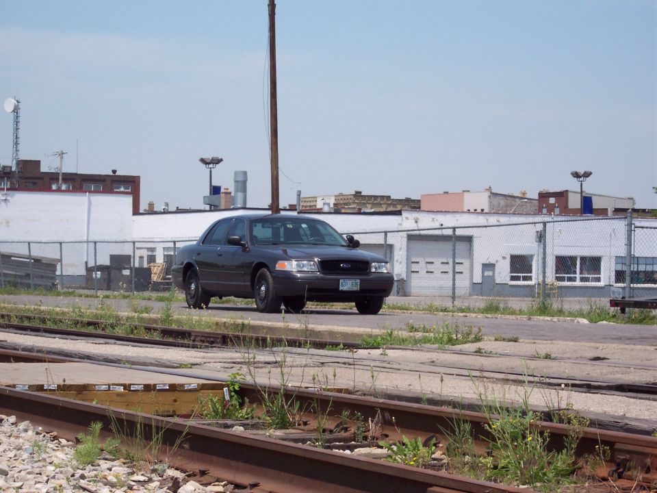 Dark sedan, possibly a 2001 Ford Crown Victoria, parked near industrial buildings and train tracks