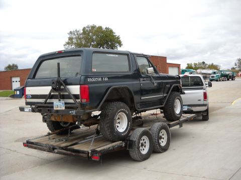 1900 Ford C-350 (Centurion Conversions) - A dark blue Ford Bronco is being transported on a trailer, with a silver pickup truck towing it. Several tanker trucks are visible in the b…