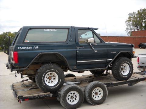 1900 Ford C-350 (Centurion Conversions) - Dark green Ford Bronco with off-road tires is chained to a trailer.