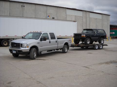 1900 Ford C-350 (Centurion Conversions) - Silver Ford F-250 tows a black Ford Bronco on a trailer, parked in front of a warehouse.